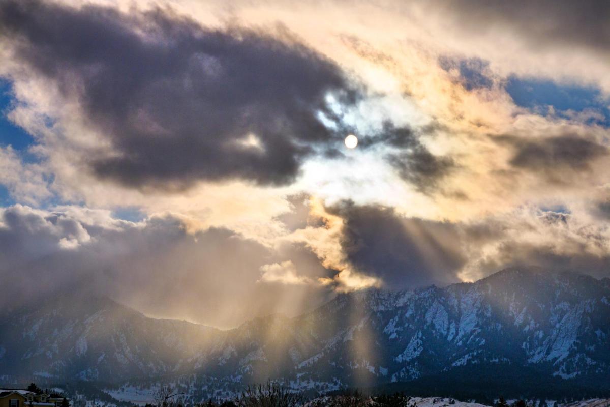Sun peaks through clouds and snowy Flatirons