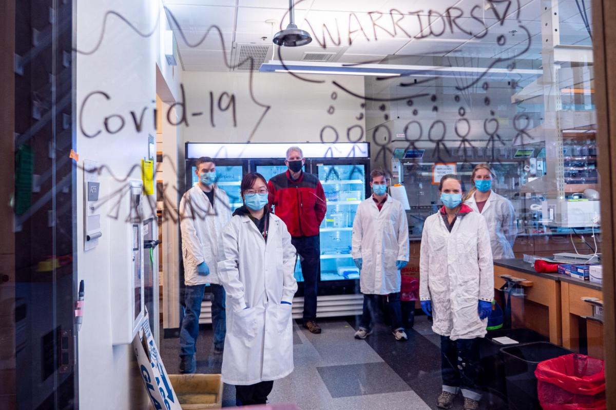 Lab members stand behind glass with the words "COVID Warriors" written on it in marker