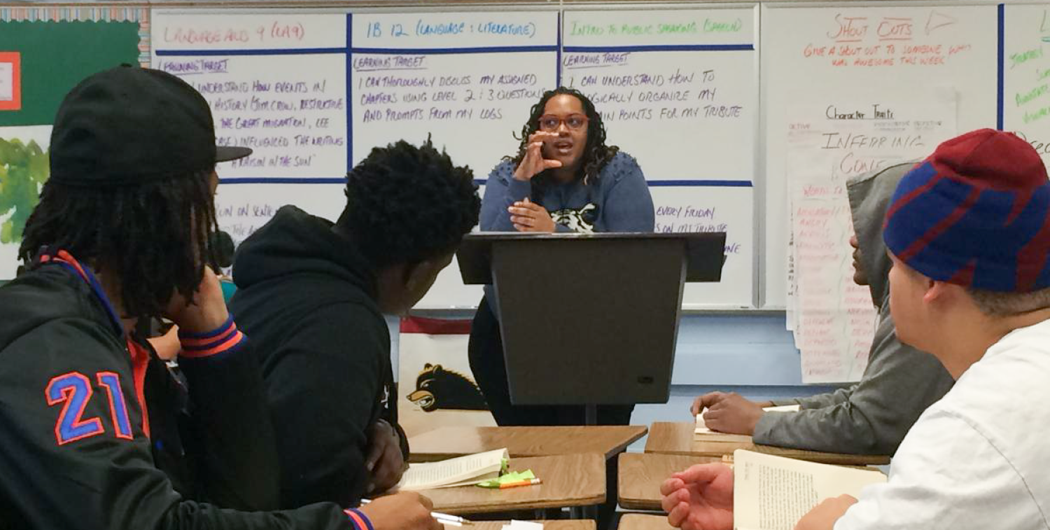 A teacher at a lectern speaks to students sitting at desks