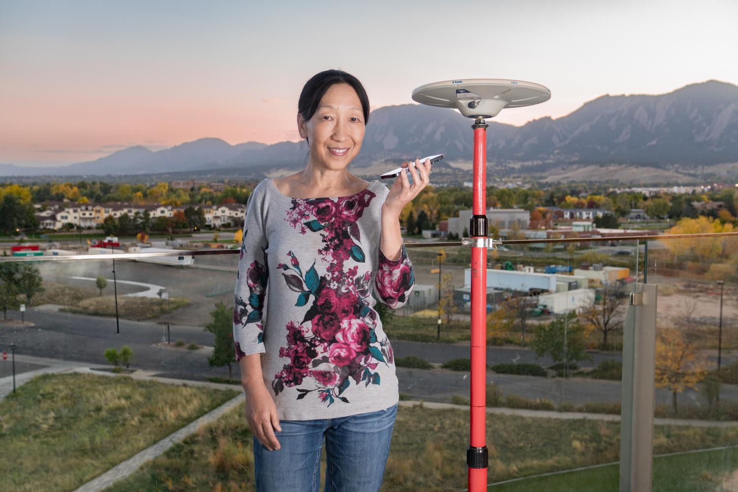 Woman standing on a balcony of a building with the Flatiron Mountains in the background. She is holding a cellphone and standing next to a small radar dish