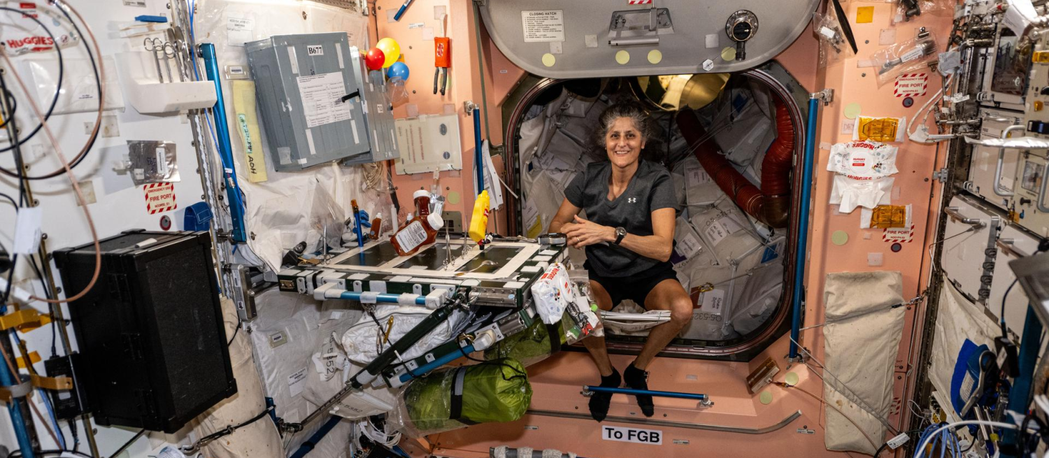 Woman wearing a t-shirt and shorts seated in a cluttered cabin aboard a space station