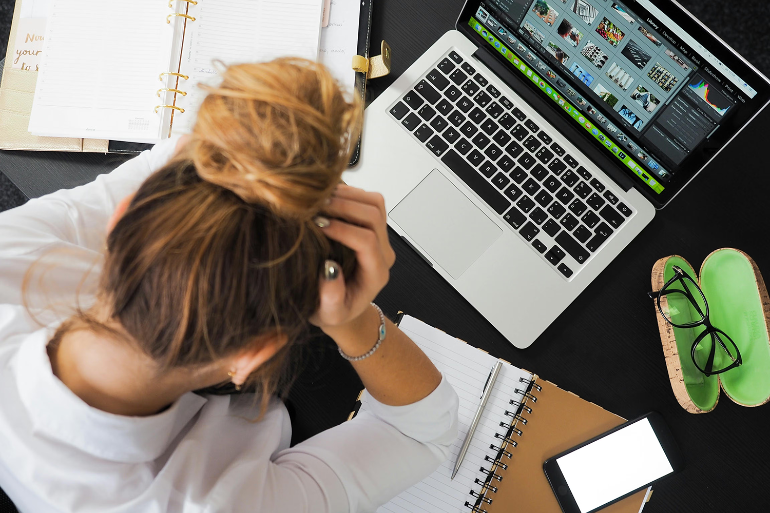 Woman seen from above at desk holding her head with a laptop and smartphone next to her