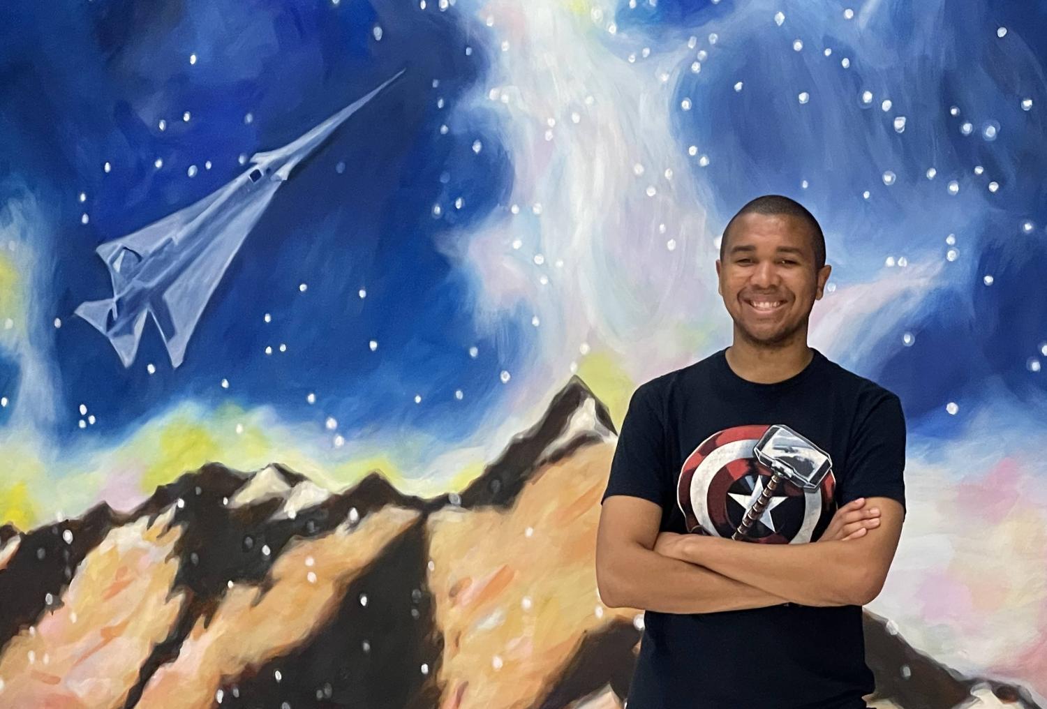 Dezell Turner stands with his arms folded in front of a mural showing mountains, a starry sky and a spaceshift flying above