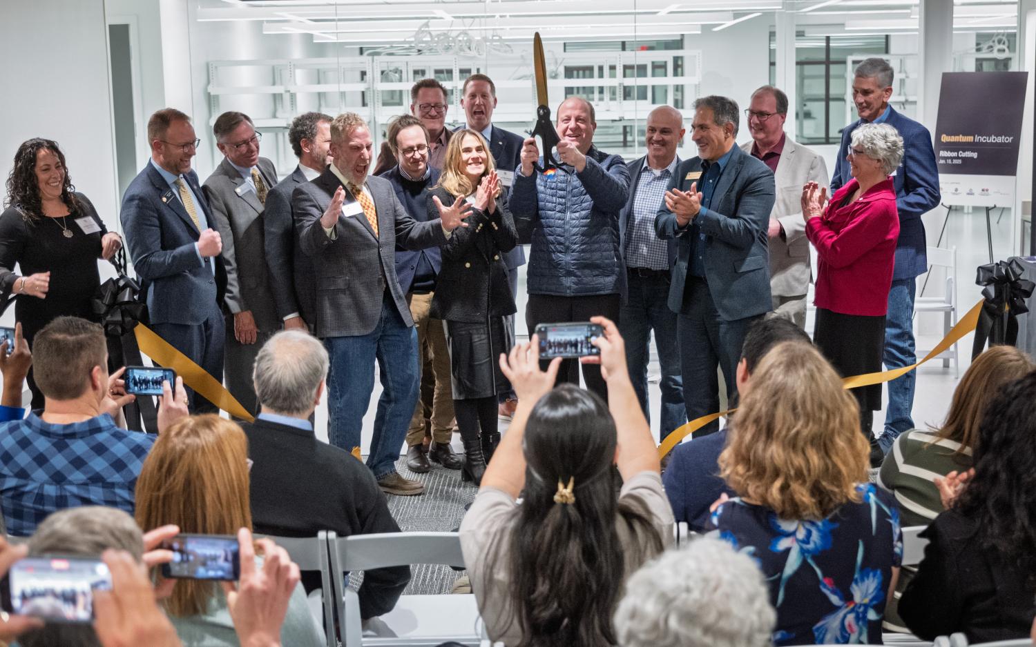 Platform guests and dignitaries watch Gov. Jared Polis celebrate after cutting a ribbon to signify the opening of the Quantum Incubator site