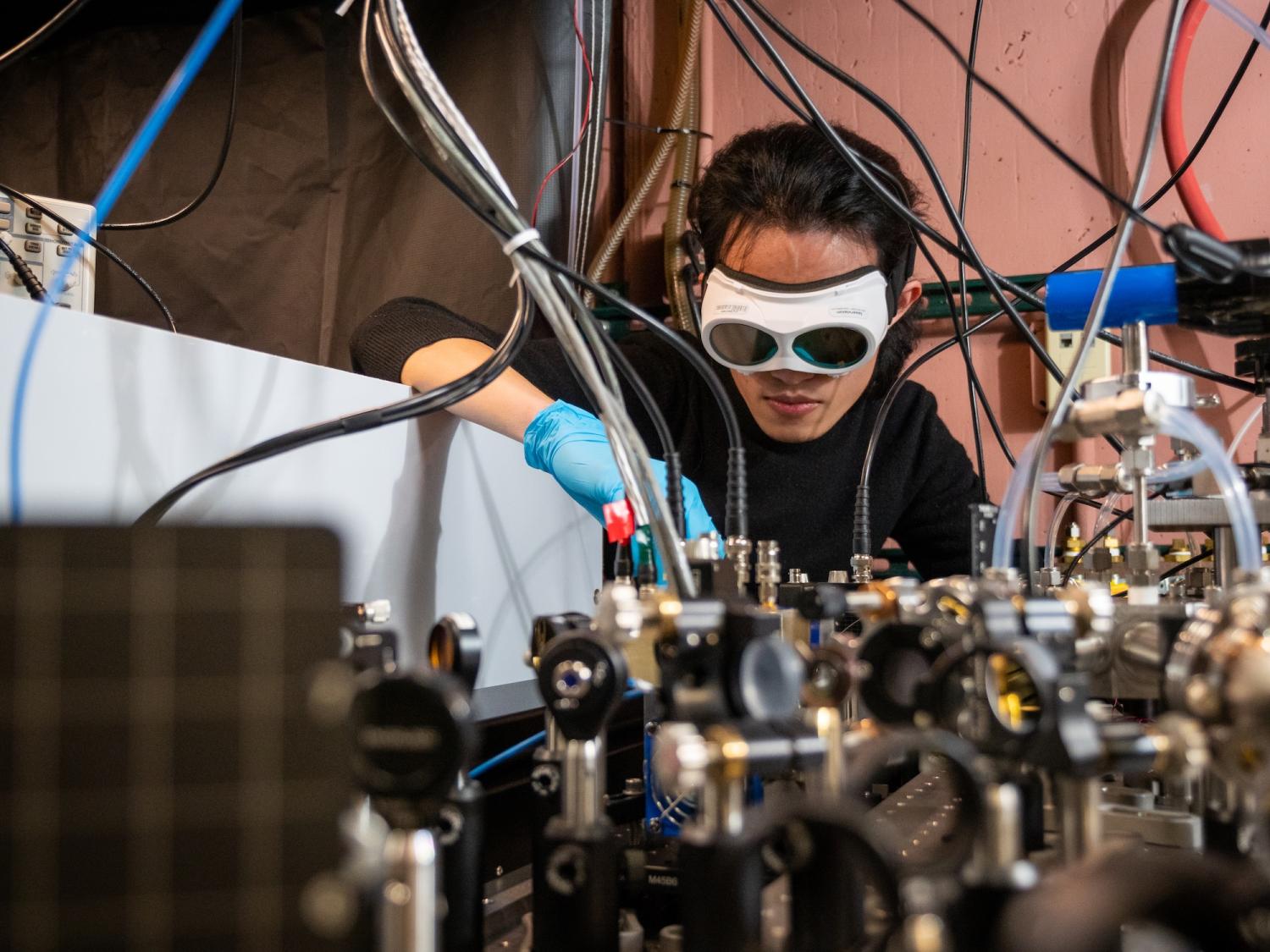 Man wearing goggles and blue gloves leans over a table holding a scientific apparatus