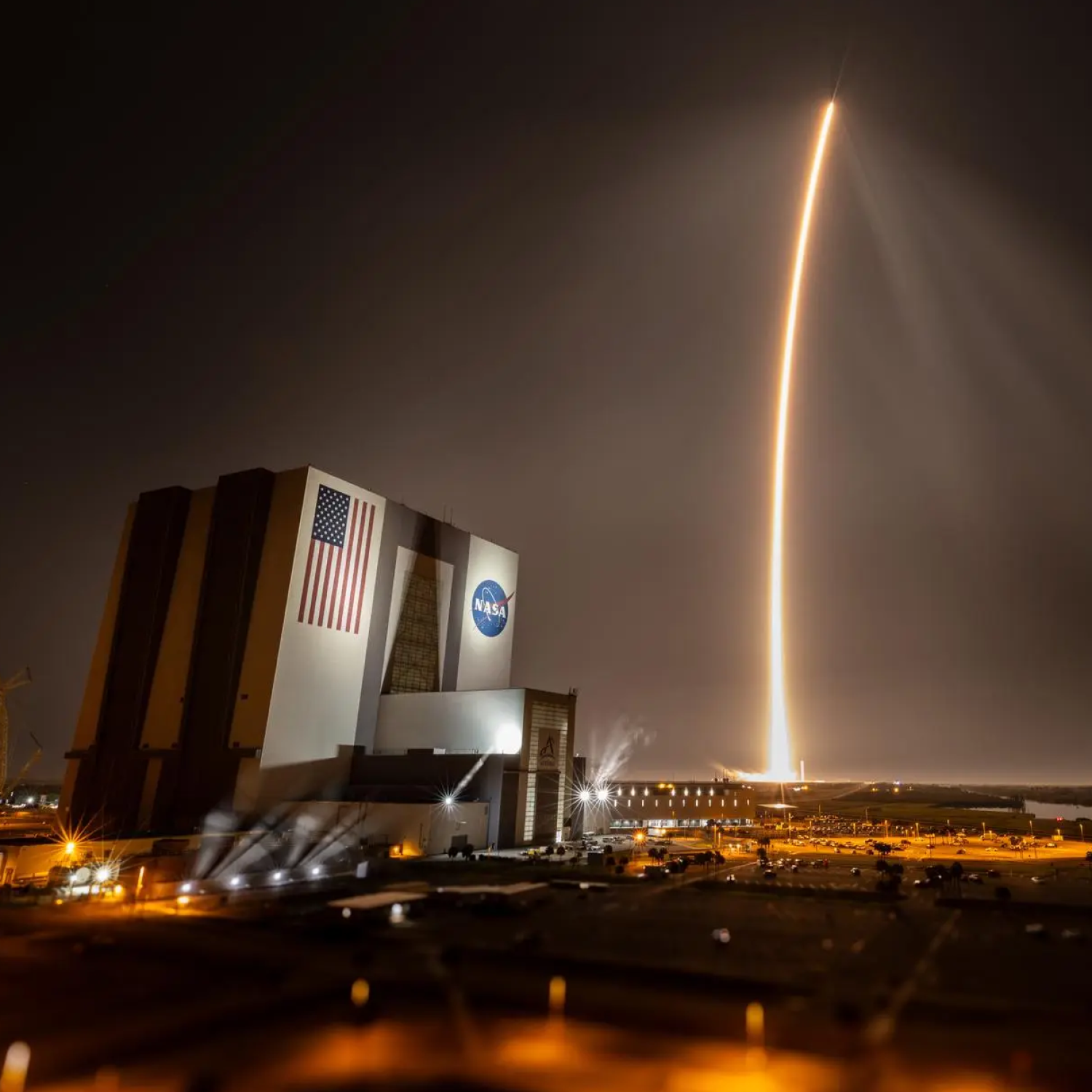 Large building showing an America flag and the NASA logo with an arc of yellow light in the background from a rocket launch