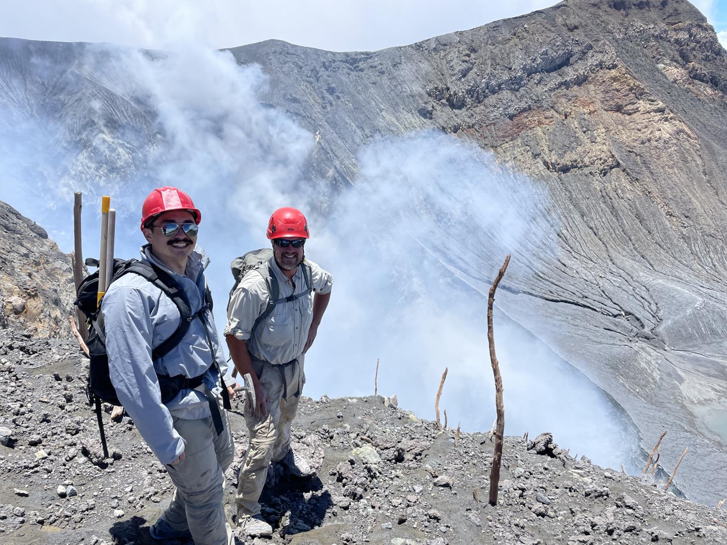 Two men wearing hard hats pose at the lip of a volcano with gas rising up behind them
