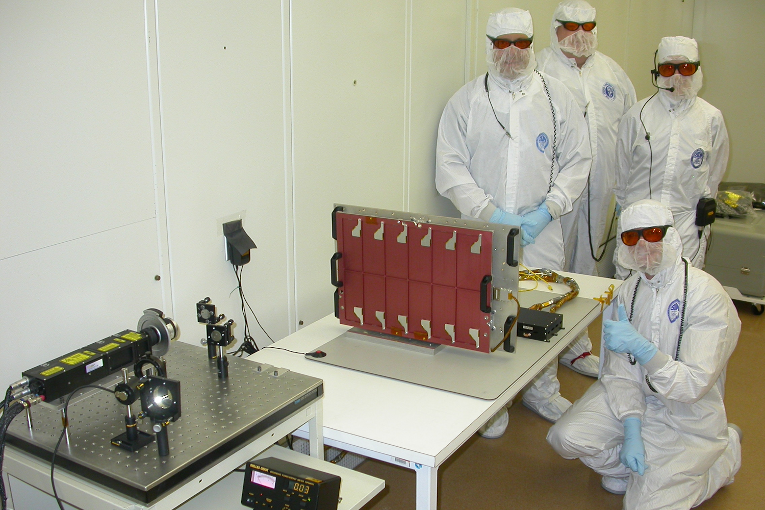 Four people wearing clean room gear posing with scientific equipment