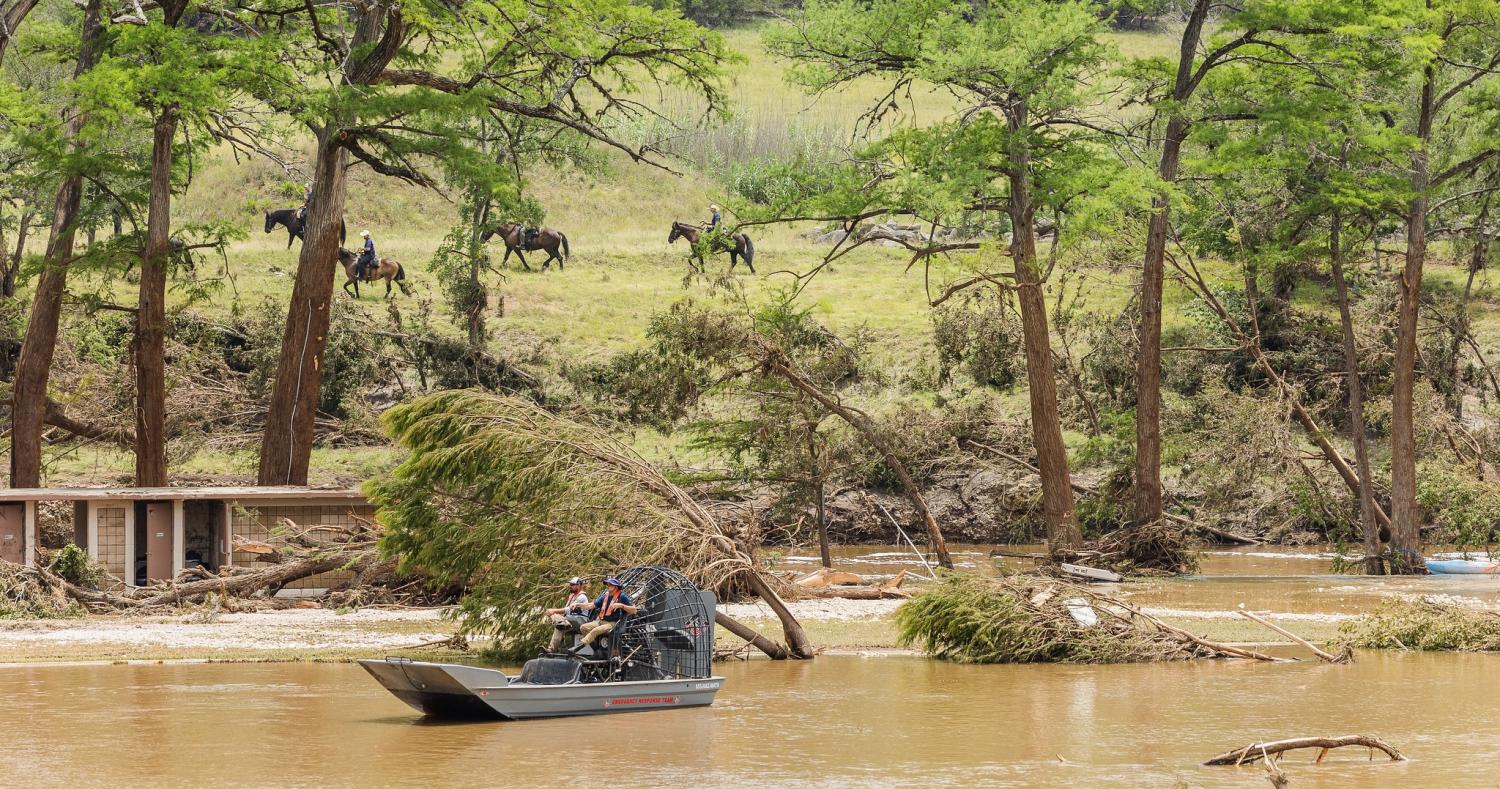 Air boat moves down swollen river with people riding horses in the background