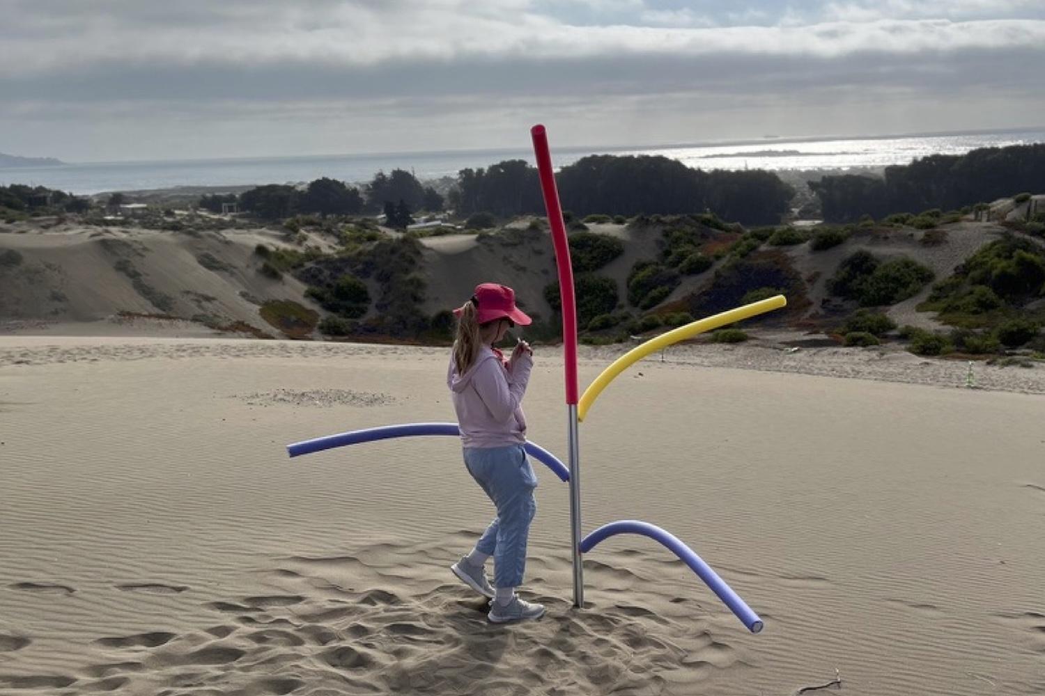 Woman on a sand dune with a metal pole sticking up with arms made from pool noodles