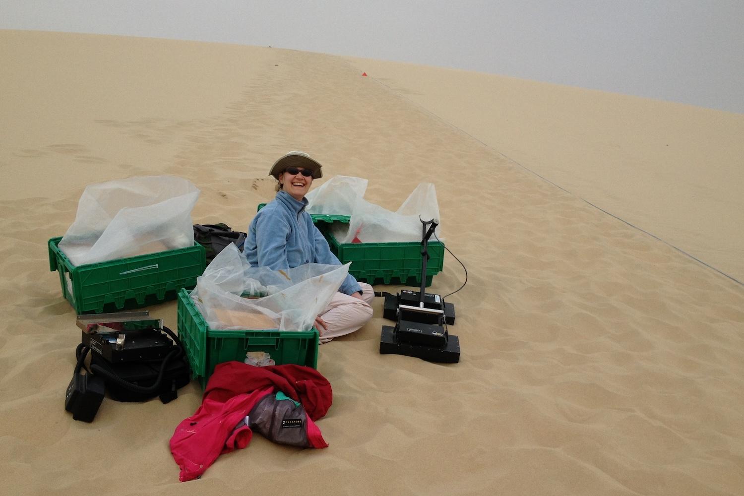 Woman sits on slope of dune surrounded by crates filled with sand
