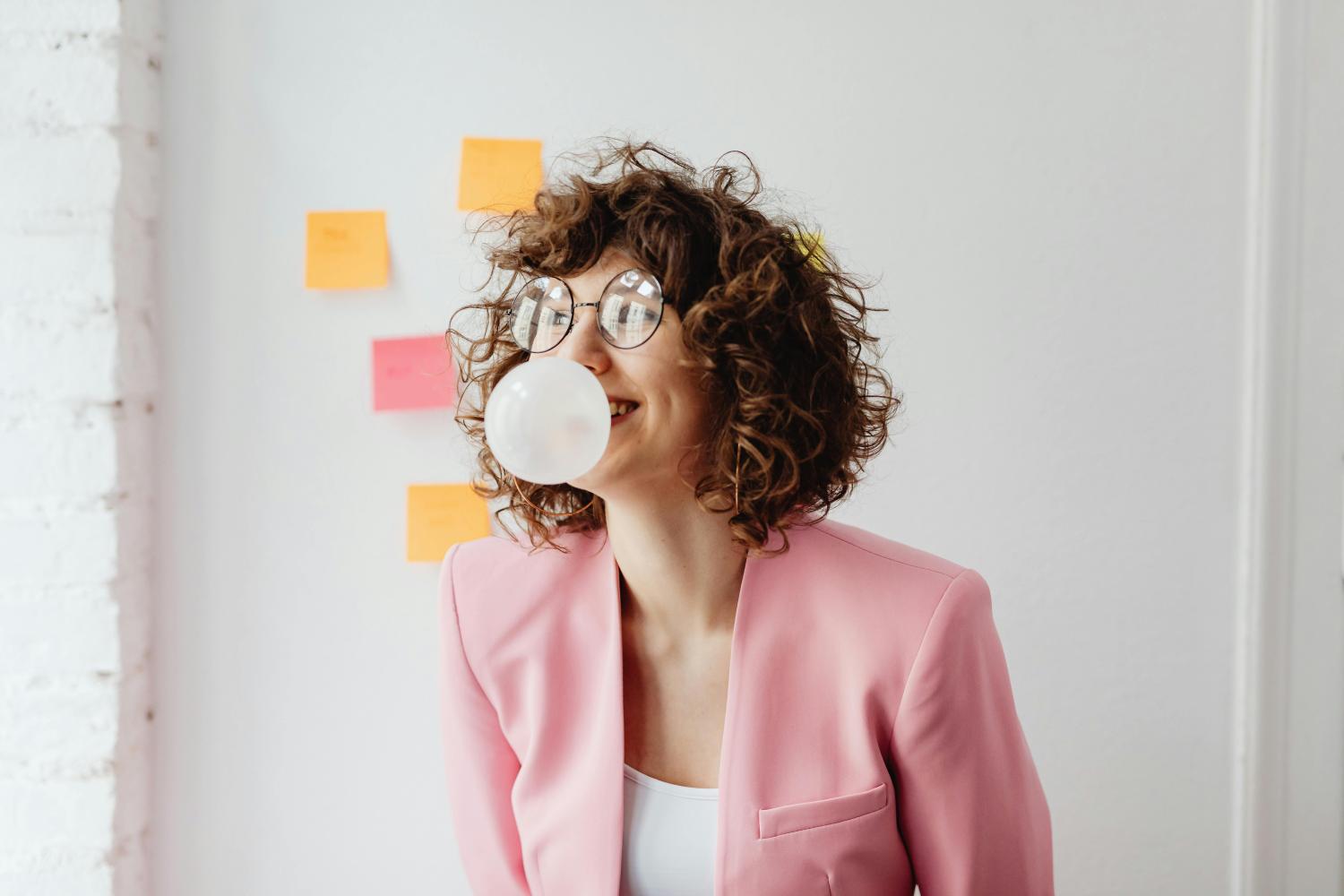 Woman blowing a bubble with gum near a board with sticky notes.