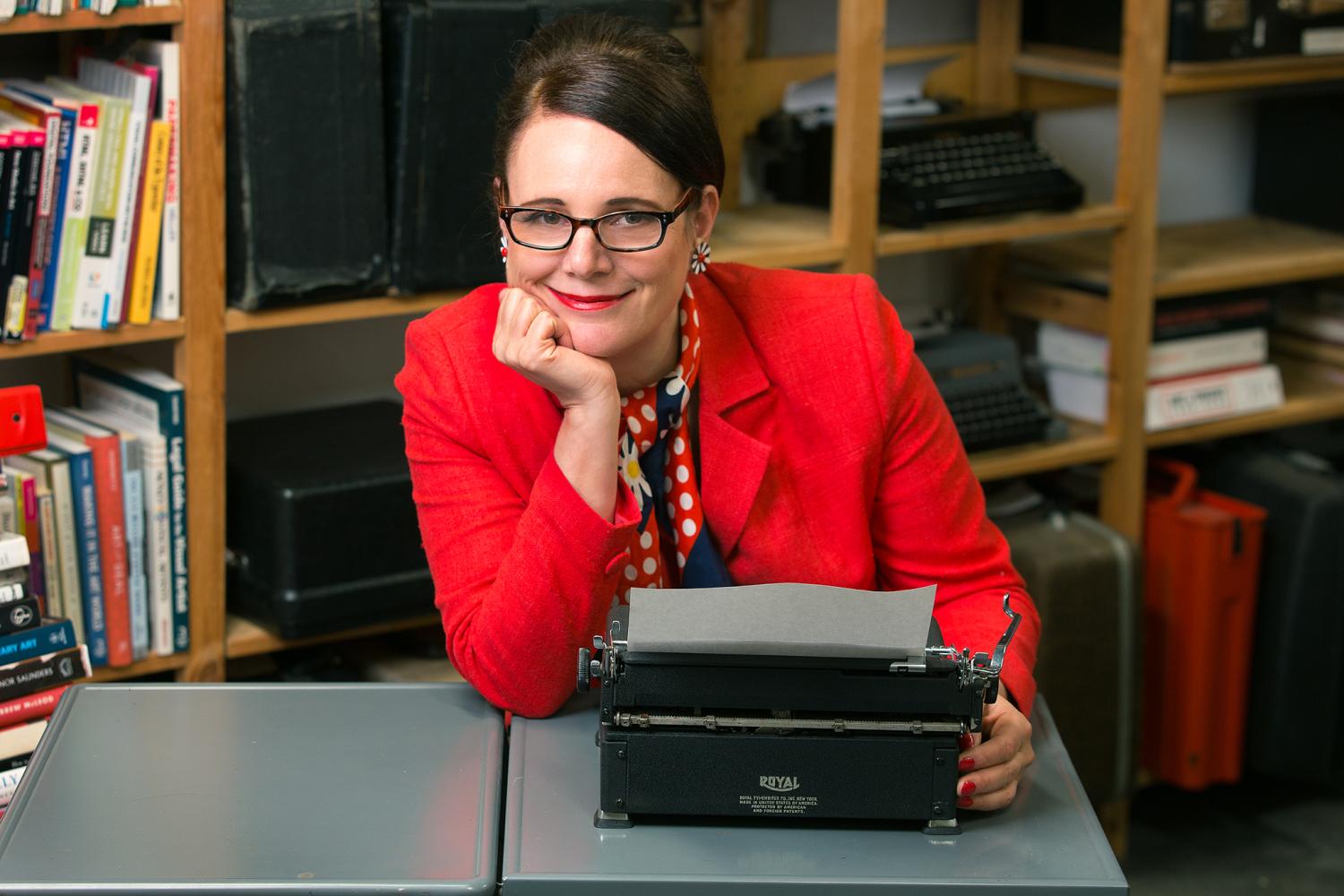 Artist Sheryl Oring, dressed in red white and blue, sits at a desk in front of her vintage typewriters.