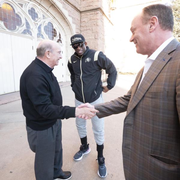 Chancellor Philip DiStefano and Athletic Director Rick George shake hands outside Macky Auditorium with Coach Prime looking on. (Casey A. Cass/University of Colorado)