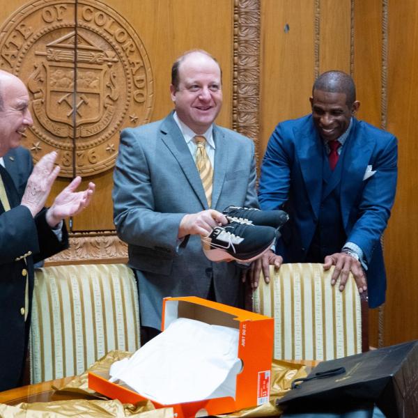 Gov. Jared Polis opens a box of CU Buffalo-branded shoes as Todd Saliman, Phil DiStefano, Deion Sanders and Wanda James look on