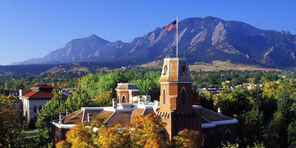 A photo of the Old Main tower with a U.S. flag on top, surrounded by colorful tree leaves and the Flatirons.