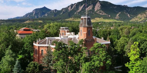 Old Main is seen in an aerial shot, surrounded by the green leaves of summer and the Flatirons in the background. (Casey Cass)