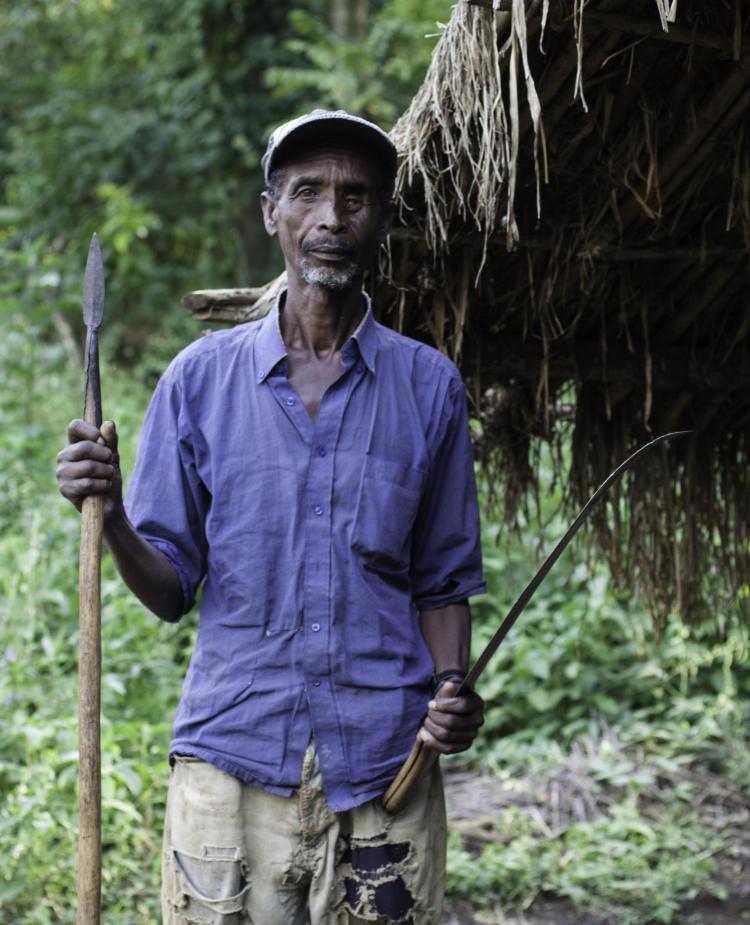 An older man holding a spear with trees in the background
