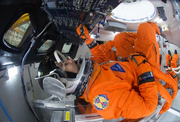 Ludzie w pomarańczowych garniturach i hełmach siedzą w ciasnym kokpicie statku kosmicznego