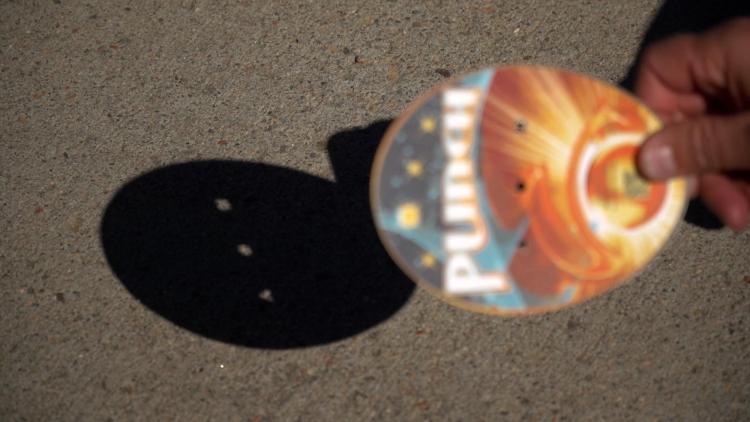 Light streams through a pinhole card, creating a projection of the sun on the sidewalk