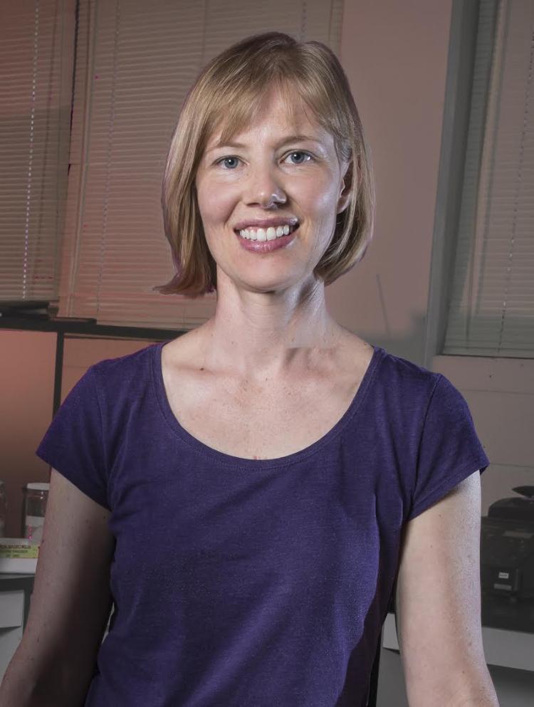 Portrait of Stacey Smith is in a purple shirt in front  of some windows with blinds in front of them.