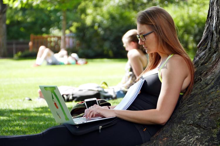 Student sits outside, leaning against tree, working on laptop