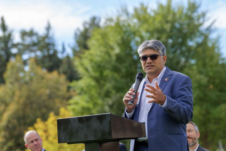 Man wearing sunglasses speaks into a microphone at a podium outdoors