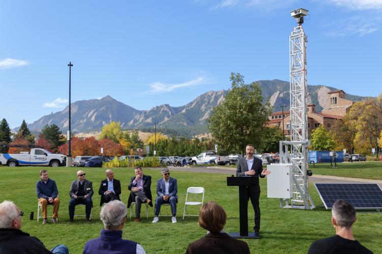 On a grassy lawn, a man speaks at a podium with several others sitting in chairs beside in. In the background, a metal tower reaches up more than 20 feet