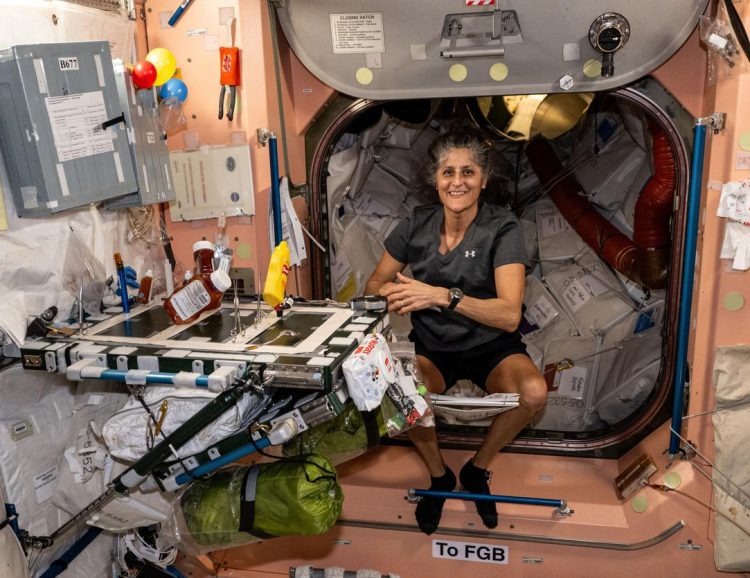 Woman wearing a t-shirt and shorts seated in a cluttered cabin aboard a space station