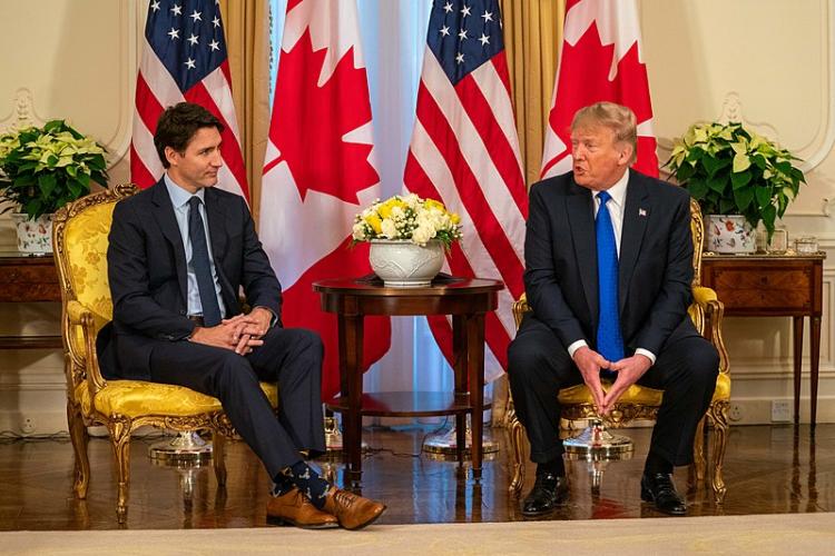 Two men, Justin Trudeau and Donald Trump, sitting on chairs in front of Canadian and American flags.