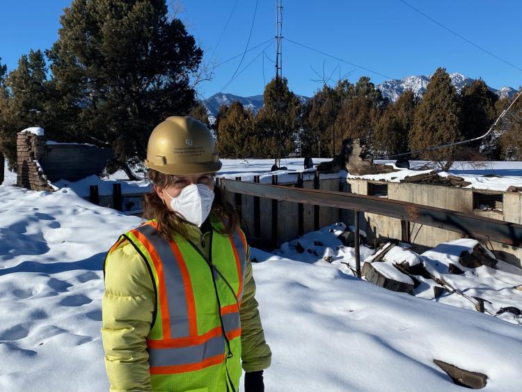 A person in a hard hat and neon vest poses for a photo with rubble from the Marshall Fire in the background.