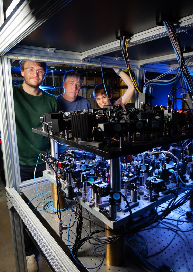 Three people stand behind a complex scientific apparatus that is glowing blue