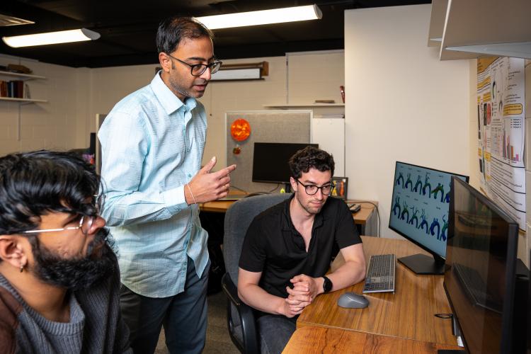 A man with a blue shirt talks with two other men sitting at a computer desk.