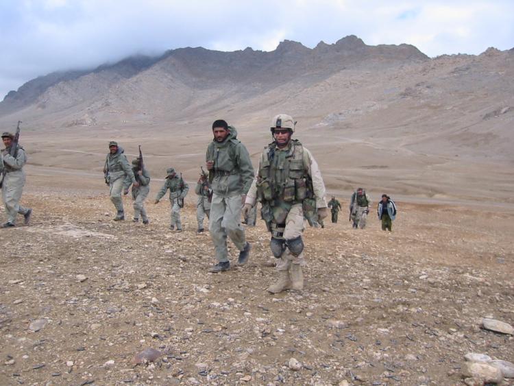 Mitch Utterback along with several other members of armed forces walk in what appears to be a dry climate, out in open space with mountains in the background.
