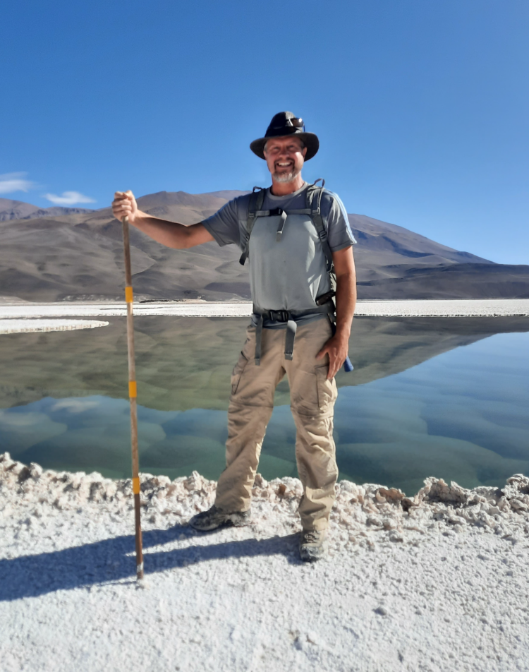 Brian Hynek holding a walking stick on the shore of a lagoon