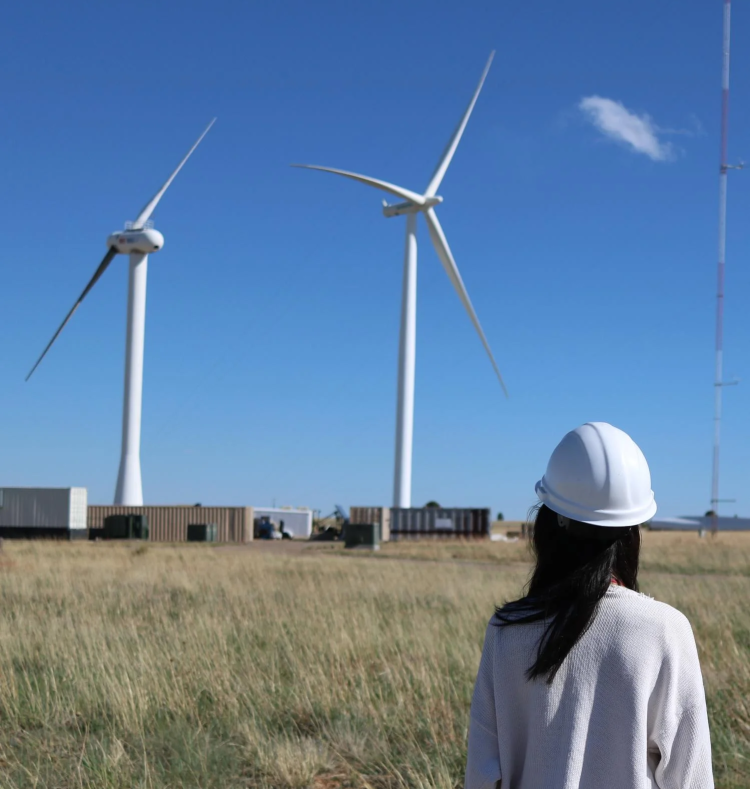 Woman wearing a hard hat stands in a field with two wind turbines in the background