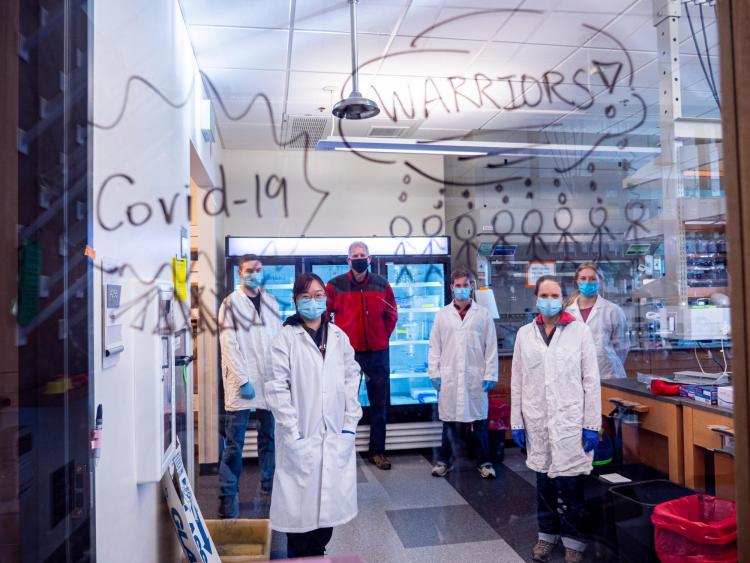 Lab members stand behind glass with the words "COVID Warriors" written on it in marker