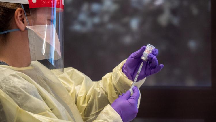 Nurse wearing a face shield and mask draws a COVID-19 vaccine out of a vial using a syring.