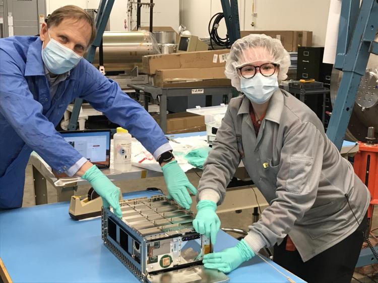 Man and woman wearing masks stand over a spacecraft about the size of a large cereal box