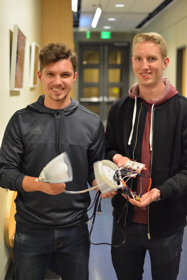 Mike Gough and Chandler Zastrow hold their Lampalarm invention in front of a hallway with photos on the wall They are mostly dressed in black.