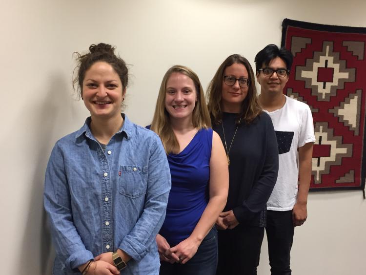 Three students and Carla Fredericks, all involved in the project, stand in a row, posing for a snapshot with a red tribal tapestry handing on the wall behind them.