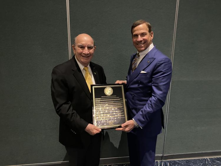 Ken Montera on the right, presents Philip DiStefano, on the left, with a chancellor emeritus plaque designed with a black background, gold writing and a gold university seal.