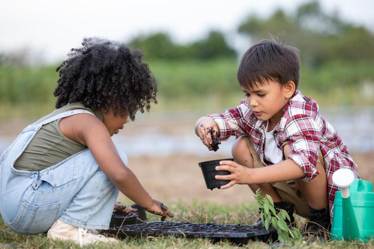 Two children kneel in the grass and scoop dirt into gardening pots