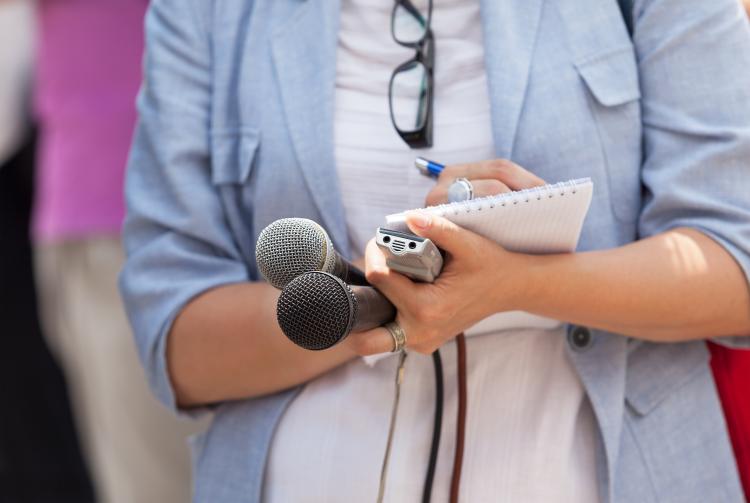 In a stock image, a journalist stands with two microphones, a reporter's notebook and pen.