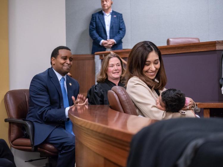 U.S. Representative Joe Neguse and Colorado Supreme Court justice Melissa Hart, center, and Andrea Neguse holding a baby listen to Governor Jared Polis speak. Photo by Glenn Asakawa.