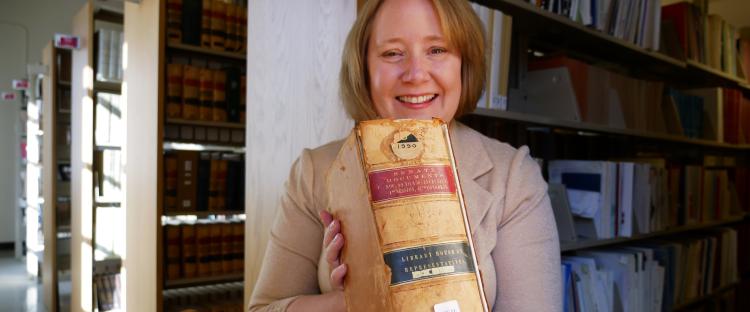 Kate Tallman is holding a thick volume of congressional records from the 1800s with bookshelves filled with books behind her in Norlin Library.