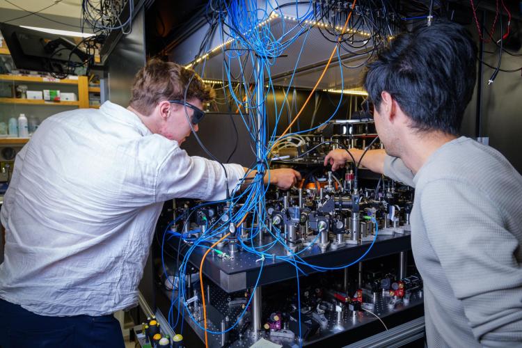 Two men seen from behind lean over a lab table that's covered in machinery, lenses and wires