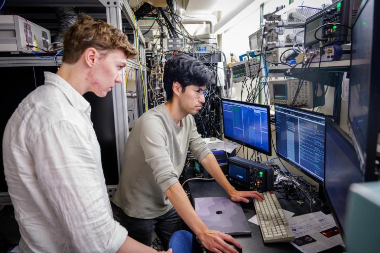 Two men stand over a desk looking at two computer monitors
