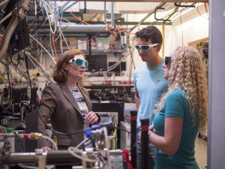 Three people wearing safety goggles talk in a lab amid scientific equipment