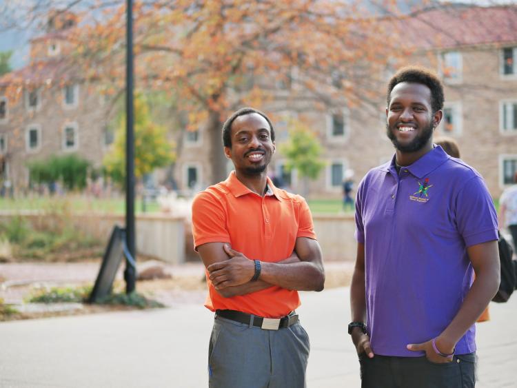 Yohannese Gebremedhin, right, and Arthur Antoine, left, stand in front of a tree and sandstone buildings in a courtyard on the University of Colorado campus.